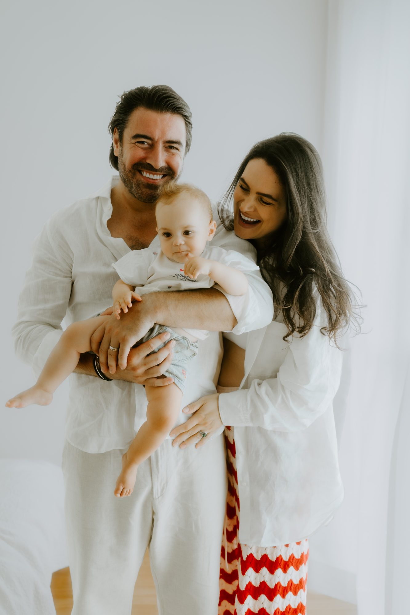 Mutter, Vater und Baby lachen gemeinsam in die Kamera, warmer Familienmoment im Licht